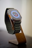 An elegant smartwatch with a rectangular display featuring a detailed digital dial. The watch has a gray metallic frame with an orange button on the side and a textured dark green strap. It rests on a stylish brown stand with a modern design.