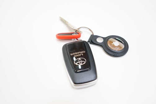 A set of keys is prominently displayed, featuring a black car key with a Land Cruiser logo and a metallic ring. Attached to the ring is a red and black keychain that holds a small circular device with what appears to be an Apple logo.