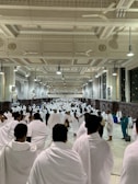 A large group of people wearing white robes walk through a spacious, ornately decorated hall with high ceilings and large ceiling fans. The architecture features columns, intricate molding, and lighting fixtures that illuminate the interior. The floor is tiled, and there are additional people wearing teal uniforms visible on the right side.