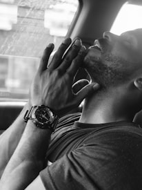 A person with eyes closed and hands clasped near their face, wearing a wristwatch, appears to be in a reflective or meditative state. The black and white filter adds a contemplative and calm atmosphere.