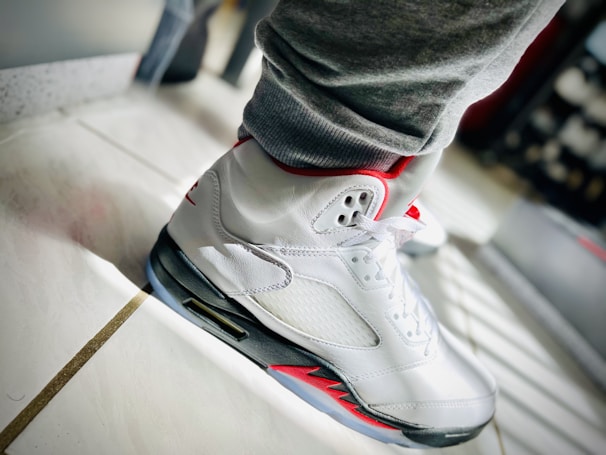 A close-up view of a white athletic sneaker with red accents is shown on a tiled floor. The shoe is laced up and the light creates a bright reflection on the white surface, highlighting its design features. The sneaker is paired with gray sweatpants, and the overall setting suggests a casual, sporty vibe.
