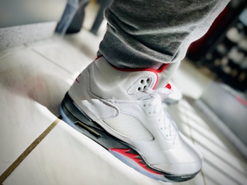 A close-up view of a white athletic sneaker with red accents is shown on a tiled floor. The shoe is laced up and the light creates a bright reflection on the white surface, highlighting its design features. The sneaker is paired with gray sweatpants, and the overall setting suggests a casual, sporty vibe.