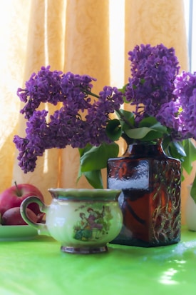 A bouquet of purple lilac flowers is arranged in an ornate dark glass vase, sitting on a green surface. Next to the vase, there is a decorative ceramic cup with artistic figures, and two red apples rest on a green plate. A warm, yellow curtain hangs in the background, allowing natural light to softly illuminate the scene.