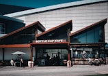 A cafe named Bottle Cap Café is set against a modern building with distinct architectural lines. The establishment features large glass windows and a slanted roof. There are a few tables and chairs outside with people enjoying their time. An umbrella provides shade for one of the tables. The cafe looks vibrant and welcoming.