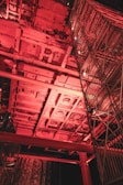 A complex industrial structure with metal frameworks and scaffolding illuminated by red lighting. The scene captures an intricate network of beams and platforms, possibly part of a construction or maintenance site.