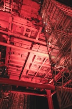 A complex industrial structure with metal frameworks and scaffolding illuminated by red lighting. The scene captures an intricate network of beams and platforms, possibly part of a construction or maintenance site.
