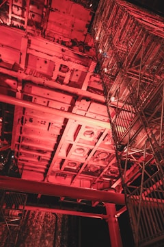 A complex industrial structure with metal frameworks and scaffolding illuminated by red lighting. The scene captures an intricate network of beams and platforms, possibly part of a construction or maintenance site.