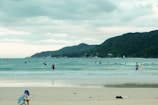 A beach scene featuring a child playing near the shore while several people swim or surf in the ocean. The sea is calm with gentle waves, and a lush green mountain range stretches across the horizon under a cloudy sky.
