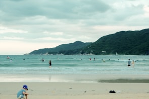 A beach scene featuring a child playing near the shore while several people swim or surf in the ocean. The sea is calm with gentle waves, and a lush green mountain range stretches across the horizon under a cloudy sky.