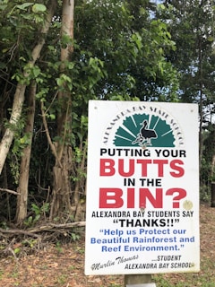 A sign in front of dense green foliage advises people to dispose of their cigarette butts properly. The text prominently reads 'PUTTING YOUR BUTTS IN THE BIN?' and highlights a message from students at Alexandra Bay School, thanking individuals for protecting the environment. A signature from a student named Murlin Thomas is visible.
