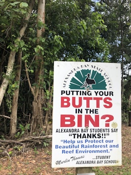 A sign in front of dense green foliage advises people to dispose of their cigarette butts properly. The text prominently reads 'PUTTING YOUR BUTTS IN THE BIN?' and highlights a message from students at Alexandra Bay School, thanking individuals for protecting the environment. A signature from a student named Murlin Thomas is visible.