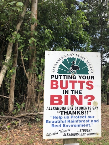 A sign in front of dense green foliage advises people to dispose of their cigarette butts properly. The text prominently reads 'PUTTING YOUR BUTTS IN THE BIN?' and highlights a message from students at Alexandra Bay School, thanking individuals for protecting the environment. A signature from a student named Murlin Thomas is visible.