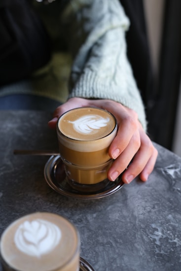 A table with a grey, textured surface displays two glasses containing creamy lattes with intricate latte art. One glass is held by a person wearing a light green, cozy sweater. The setting conveys a relaxed and warm atmosphere, ideal for enjoying a coffee break.