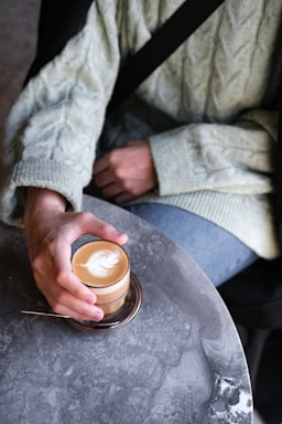 A person wearing a cozy, light-colored cable-knit sweater is sitting at a marble table. They are holding a glass cup containing a latte with latte art on top, featuring a leaf pattern.