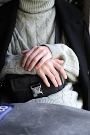 A person is wearing a soft, light grey cable-knit sweater and a black winter coat. Their hands are gently resting on a small black bag with a metal buckle.