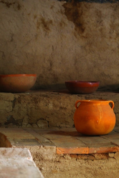 Three clay pots of varying sizes are placed on rustic stone steps, with an earthy, textured wall in the background. The pots have a natural, unglazed finish, and their muted orange and brown hues blend harmoniously with the surrounding environment.