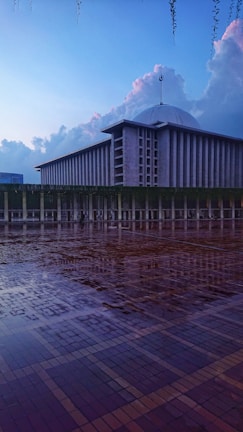 A large, modern mosque is depicted with a distinctive dome and towering minaret. The foreground features a wet, reflective tiled courtyard, enhancing the serene and tranquil atmosphere. The sky is expansive, with soft clouds catching the fading light of dusk.