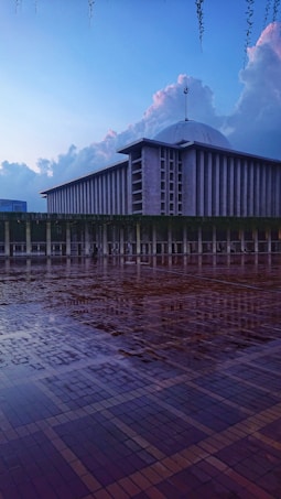 A large, modern mosque is depicted with a distinctive dome and towering minaret. The foreground features a wet, reflective tiled courtyard, enhancing the serene and tranquil atmosphere. The sky is expansive, with soft clouds catching the fading light of dusk.