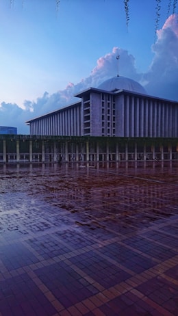 A large, modern mosque is depicted with a distinctive dome and towering minaret. The foreground features a wet, reflective tiled courtyard, enhancing the serene and tranquil atmosphere. The sky is expansive, with soft clouds catching the fading light of dusk.