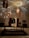 A dimly lit room with woven, rattan-style pendant lights casting intricate shadows on the ceiling. A large wooden table with ornate objects stands atop a patterned rug. Framed artwork and wooden cabinets adorn the walls, with an open doorway leading outside.