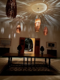 A dimly lit room with woven, rattan-style pendant lights casting intricate shadows on the ceiling. A large wooden table with ornate objects stands atop a patterned rug. Framed artwork and wooden cabinets adorn the walls, with an open doorway leading outside.