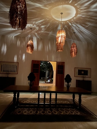 A dimly lit room with woven, rattan-style pendant lights casting intricate shadows on the ceiling. A large wooden table with ornate objects stands atop a patterned rug. Framed artwork and wooden cabinets adorn the walls, with an open doorway leading outside.