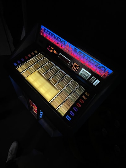 An illuminated vintage jukebox with a brightly lit display showing rows of song selections. The top part has the words 'VIDEO MUSIC' in neon blue and red with a soundwave graphic. The overall setting is dimly lit, highlighting the glow from the jukebox.