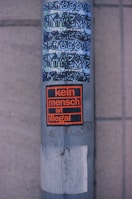 A metallic pole with several stickers is seen. The top stickers display graffiti-style text in a mix of blue and black, while the lower sticker, framed in orange and black, reads 'kein mensch ist illegal.' The base of the pole appears worn and weathered, featuring some peeled-off remnants of old stickers.