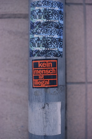 A metallic pole with several stickers is seen. The top stickers display graffiti-style text in a mix of blue and black, while the lower sticker, framed in orange and black, reads 'kein mensch ist illegal.' The base of the pole appears worn and weathered, featuring some peeled-off remnants of old stickers.
