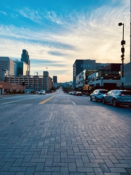 A city street stretches into the distance, lined with modern buildings on both sides. The foreground features a patterned brick road, while cars are parked along the sidewalk. The sky above is expansive, with scattered clouds illuminated by soft, warm sunlight. A prominent building displays the word 'OMAHA'.