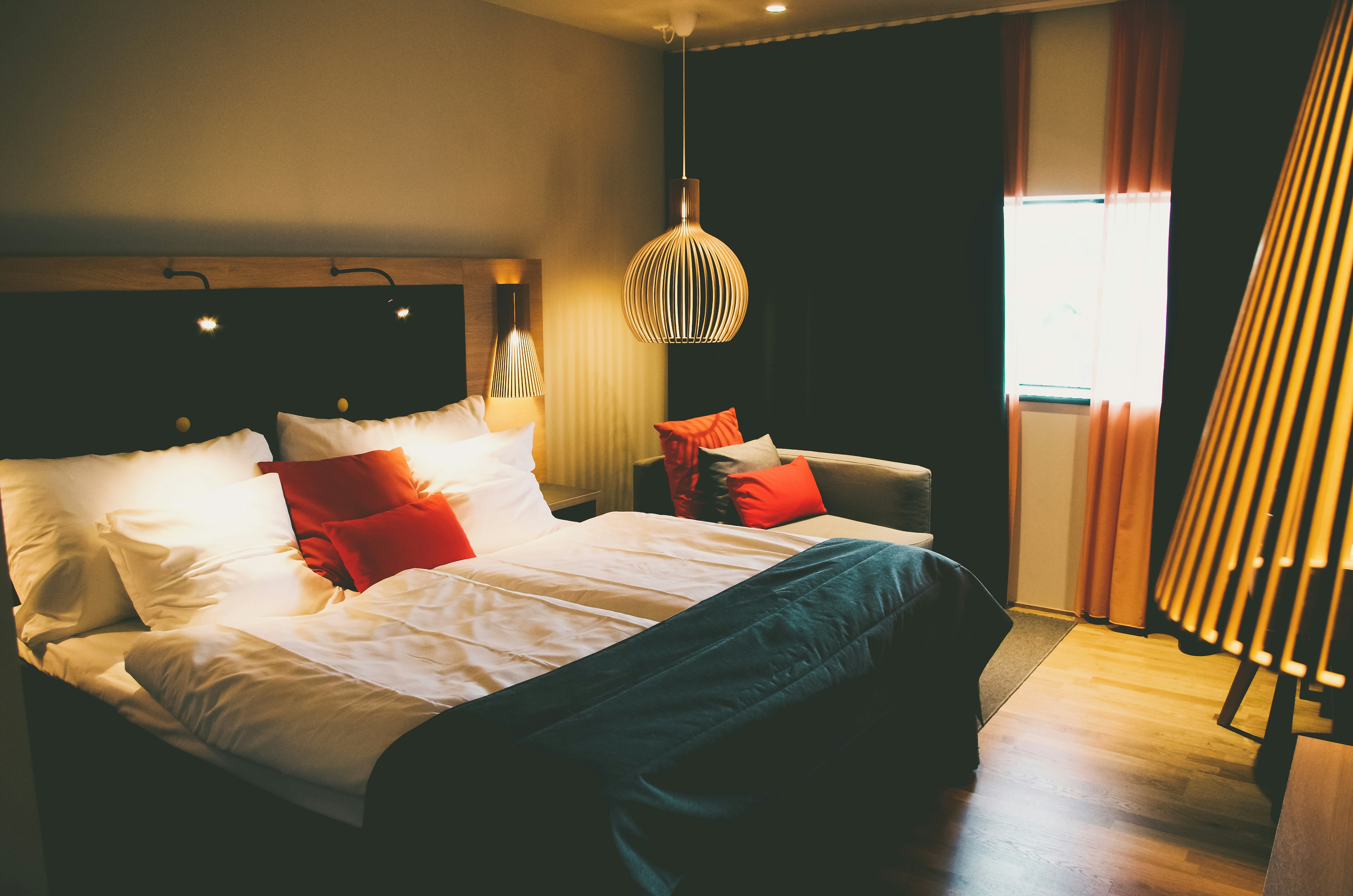 A cozy and modern hotel room features a large bed with white and red pillows, complemented by a dark bedspread. The room has ambient lighting with a unique pendant lamp above the bedside, casting warm light. On one side, a comfortable couch with more pillows sits by the window, framed by elegant red curtains. Wooden flooring and other warm-tone decor enhance the inviting atmosphere.