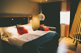 A cozy and modern hotel room features a large bed with white and red pillows, complemented by a dark bedspread. The room has ambient lighting with a unique pendant lamp above the bedside, casting warm light. On one side, a comfortable couch with more pillows sits by the window, framed by elegant red curtains. Wooden flooring and other warm-tone decor enhance the inviting atmosphere.