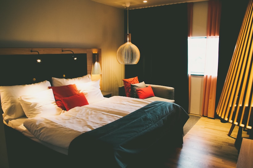 A cozy and modern hotel room features a large bed with white and red pillows, complemented by a dark bedspread. The room has ambient lighting with a unique pendant lamp above the bedside, casting warm light. On one side, a comfortable couch with more pillows sits by the window, framed by elegant red curtains. Wooden flooring and other warm-tone decor enhance the inviting atmosphere.