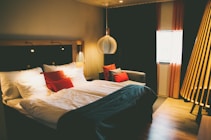 A cozy and modern hotel room features a large bed with white and red pillows, complemented by a dark bedspread. The room has ambient lighting with a unique pendant lamp above the bedside, casting warm light. On one side, a comfortable couch with more pillows sits by the window, framed by elegant red curtains. Wooden flooring and other warm-tone decor enhance the inviting atmosphere.