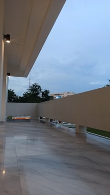 A modern architectural exterior with a smooth, polished marble floor reflecting the ambient light. The design features clean lines and a minimalistic railing, allowing a view of the outdoors with green trees and distant buildings. Soft lighting fixtures are mounted on the ceiling.