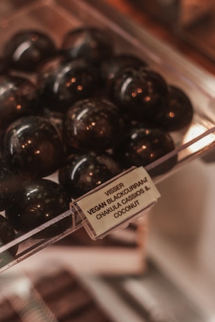 A display of glossy, dark spherical objects, possibly chocolates or candy, is set inside a clear plastic container. There is a label identifying the contents as 'Vegan Blackcurrant ChakuLa Cassios & Coconut.' The setting suggests a store or a confectionery shop.
