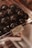 A display of glossy, dark spherical objects, possibly chocolates or candy, is set inside a clear plastic container. There is a label identifying the contents as 'Vegan Blackcurrant ChakuLa Cassios & Coconut.' The setting suggests a store or a confectionery shop.