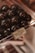 A display of glossy, dark spherical objects, possibly chocolates or candy, is set inside a clear plastic container. There is a label identifying the contents as 'Vegan Blackcurrant ChakuLa Cassios & Coconut.' The setting suggests a store or a confectionery shop.