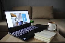 A cozy setting with a laptop on a wooden table, showing images of architecture on the screen. Nearby, there is an open notebook and a coffee mug placed on a coaster. A small potted plant adds a touch of greenery, and the background features a beige sofa.