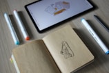 A tablet displaying a digital drawing sits on a wooden surface, surrounded by markers with different colored tips. An open notebook reveals a sketch of a sneaker. The scene suggests a workspace for design or illustration.