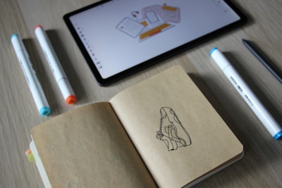 A tablet displaying a digital drawing sits on a wooden surface, surrounded by markers with different colored tips. An open notebook reveals a sketch of a sneaker. The scene suggests a workspace for design or illustration.