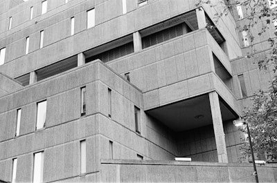 A large, multi-story brutalist building with geometric lines and concrete panels fills the frame. The building features narrow, rectangular windows and multiple layers creating a boxy, angular appearance. In the foreground, some tree branches and a lamp post are visible, adding a natural element to the urban scene.