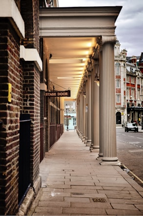 A long, covered walkway with classical columns leads to a 'Stage Door' sign. The walkway is illuminated by regular, evenly spaced lights on the ceiling. To the right, there is a view of a street with historical buildings, featuring ornate architecture with brick and stone facades.