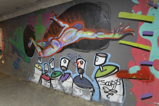 Graffiti artwork features human figures in vibrant colors. The central figure appears to be in motion, with exaggerated limbs and a rainbow-like hue. Below, abstract shapes resemble spray paint cans with faces.