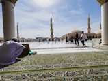 A spacious courtyard of a mosque with beautifully decorated minarets in the background. A few people in traditional attire are walking, while one person sits on a green prayer rug, engaged in reading or reflection.