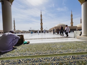 A spacious courtyard of a mosque with beautifully decorated minarets in the background. A few people in traditional attire are walking, while one person sits on a green prayer rug, engaged in reading or reflection.