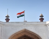 An Indian flag waves prominently atop a white historical building with two ornate turrets flanking it. The architecture features intricate carvings and a large arched entrance below. There is a clear, blue sky in the background.