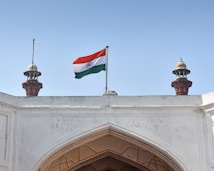 An Indian flag waves prominently atop a white historical building with two ornate turrets flanking it. The architecture features intricate carvings and a large arched entrance below. There is a clear, blue sky in the background.