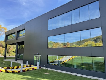 A modern, sleek building with reflective glass windows that mirror the lush green surroundings and clear blue sky. The facade is predominantly a dark gray color, complementing the vibrant landscaping of colorful flowers lining the entrance path. The setting appears peaceful and well-maintained, with a manicured lawn and a backdrop of deciduous trees in their early autumn hues.