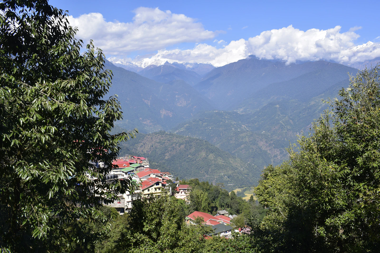 Red roofed hotels of Pelling among forest on the ridge, with the valleys and cloud capped snow peaks of West Sikkim beyond